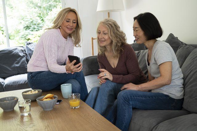 Mid-shot of middle age woman showing photos on her phone to two female friends while sitting on sofa at home