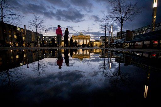 11 March 2020, Berlin: Passers-by walk across Pariser Platz at dusk in front of the Brandenburg Gate. The scenery is reflected in a puddle. Photo: Christoph Soeder\/