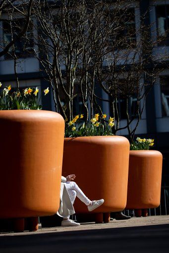 25 March 2020, Hamburg: A woman sits alone on a bench between large flower pots in the sun. Photo: Christian Charisius\/