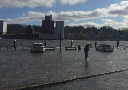 29 March 2020, Schleswig-Holstein, Flensburg: A man is standing next to cars on the flooded parking lot Schiffbrücke in the city centre, knee-deep in water. On the entire Baltic Sea coast, water levels have risen significantly as a result of the announced storm surge. In many places they were more than one metre above the mean water level. Photo: Birgitta von Gyldenfeldt\/
