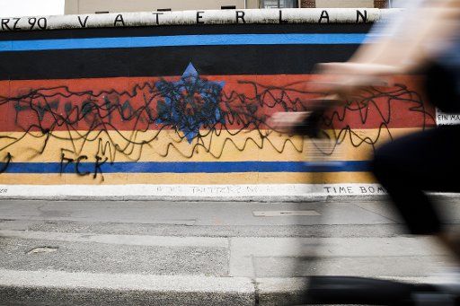 22 May 2020, Berlin: A cyclist rides past the destroyed wall painting by the artist Günther Schäfer at the East Side Gallery. The mural "Vaterland", which shows the black-red-golden German flag with a blue Star of David, was smeared with black graffiti by unknown persons. Photo: Carsten Koall\/