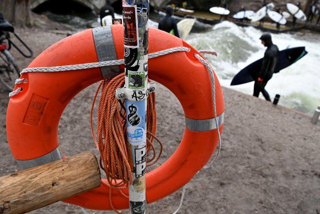 18 March 2021, Bavaria, Munich: An orange life preserver hangs on the Eisbach, while surfers ride the artificial wave in the background. Photo: Peter Kneffel\/dpa
