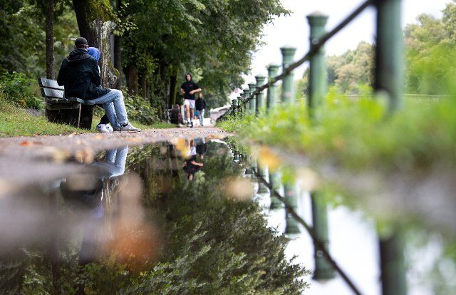 31 August 2021, Bavaria, Munich: A man and a woman are sitting on a bench by the Nymphenburg Canal, reflecting in a puddle. Photo: Sven Hoppe\/dpa\/ZB