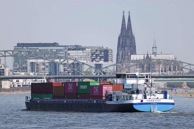 18 May 2022, North Rhine-Westphalia, Cologne: A barge, loaded with containers, sails across the Rhine in Cologne in front of the cathedral (r) and the crane houses (l) Photo: Oliver Berg\/dpa