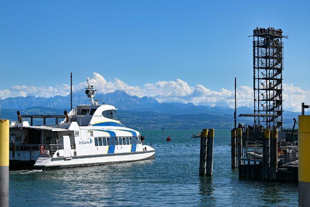 25 June 2022, Baden-Wuerttemberg, Friedrichshafen Am Bodensee: The catamaran Ferdinand sails out of the harbor while dozens of people stand on and around the Moleturm. The Alps in Switzerland can be seen in the background. Photo: Felix Kästle\/dpa