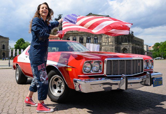 31 May 2022, Saxony, Dresden: Daniela Hesse, project manager of the US Car Convention 2022, stands with a US flag at a press event next to a Ford Torino, known from the movie Starsky & Hutch, in front of the Semper Opera House on Theaterplatz. Around 2,000 vehicles built in the U.S. are expected at the tradition-rich summer festival in Dresden from July 8 to 10, 2022. Photo: Robert Michael\/dpa
