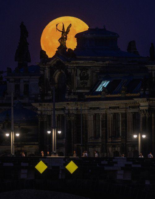 11 August 2022, Saxony, Dresden: The moon rises in the evening behind the "Eros" on the eastern corner risalit of the Academy of Fine Arts while people walk across the Augustus Bridge. On August 12, 2022, the full moon will once again become the so-called supermoon. Photo: Robert Michael\/dpa