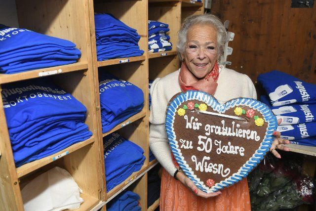 26 September 2022, Bavaria, Munich: Entrepreneur Rosi Deutsch stands in a souvenir stand in the Augustiner tent with a Wiesn heart with the inscription "We congratulate you on 50 years at the Wiesn". Deutsch is celebrating 50 years of working at the Oktoberfest this year. The Wiesn will be held from Sept. 17 to Oct. 3, 2022. Photo: Felix Hörhager\/dpa