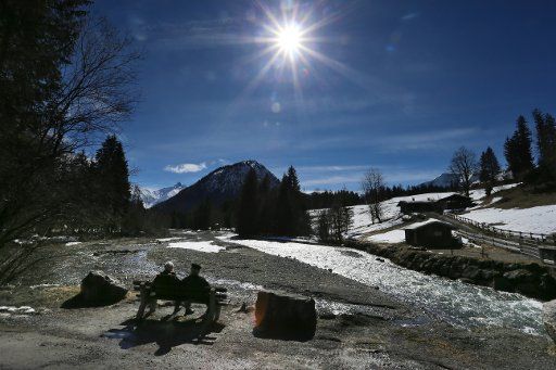 16 March 2019, Bavaria, Oberstdorf: Tourists sit on a bench at the Trettach in front of snow-covered mountains in the sunshine. Photo: Karl-Josef Hildenbrand\/