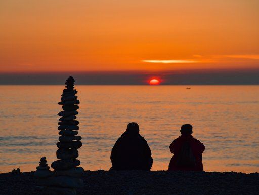 21 April 2019, Denmark, Klitmöller: Next to a stone tower, two people sit on the beach and enjoy the colourful sunset over the North Sea. Photo: Patrick Pleul\/dpa-Zentralbild\/