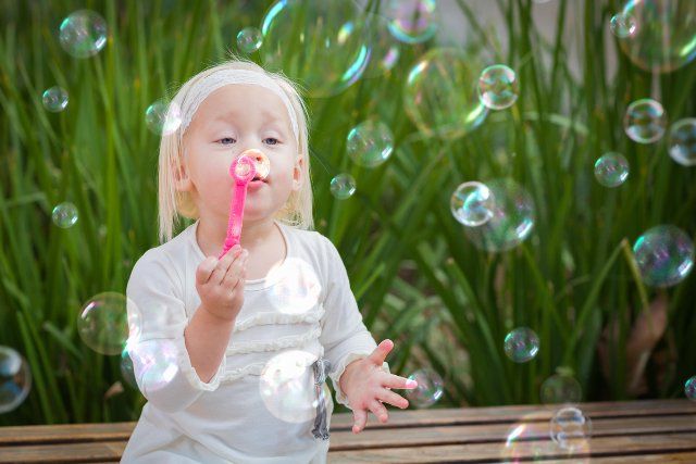 Adorable little girl sitting on bench having fun with blowing bubbles