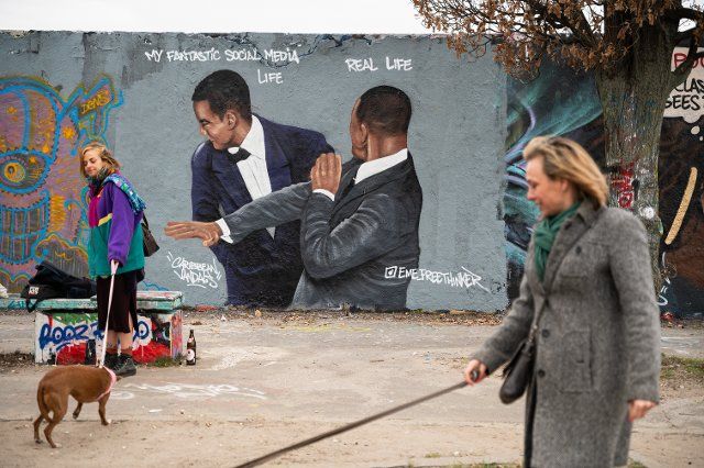 March 30, 2022 - Berlin, Germany, Europe: Two women walk their dogs in front of a graffiti by the artist Eme Freethinker on a segment of the Berlin Wall at Berliner Mauerpark in the borough of Prenzlauer Berg that depicts the actor Will Smith slapping the comedian Chris Rock in the face on stage during the 94th Academy Awards ceremony, after the comic made a joke about the actor\